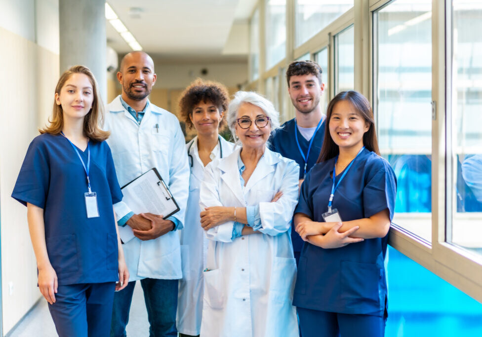 Portrait featuring a multi-ethnic medical team smiling confidently at the camera while gathered in a bustling hospital corridor